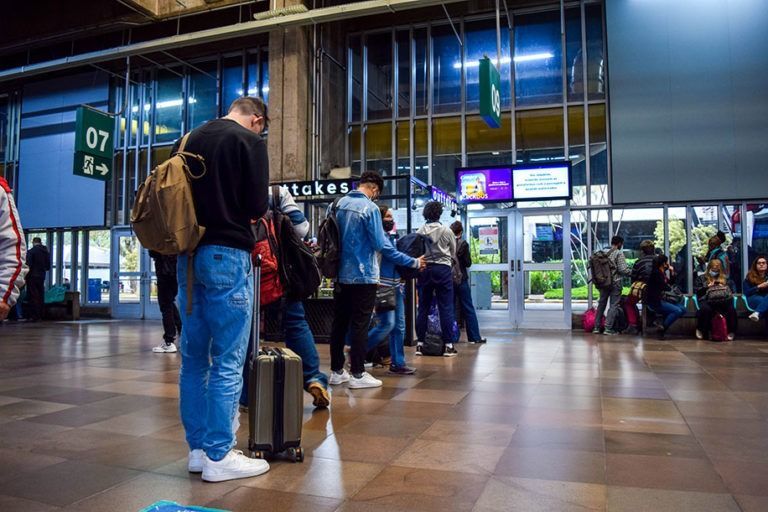 Pessoas fazem fila dentro de uma estação com bagagens. Uma pessoa verifica um celular.