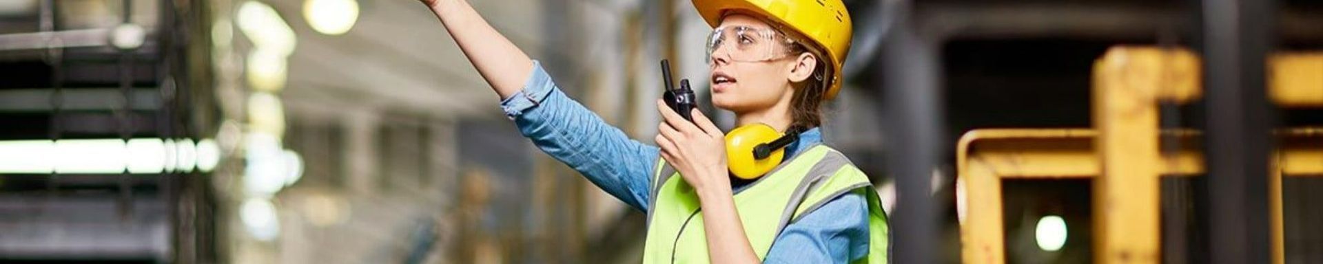 A woman wearing a hard hat and safety vest is talking on a walkie talkie in a factory.