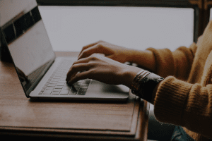 Hands typing on a laptop at a wooden table, with a brown sweater visible.