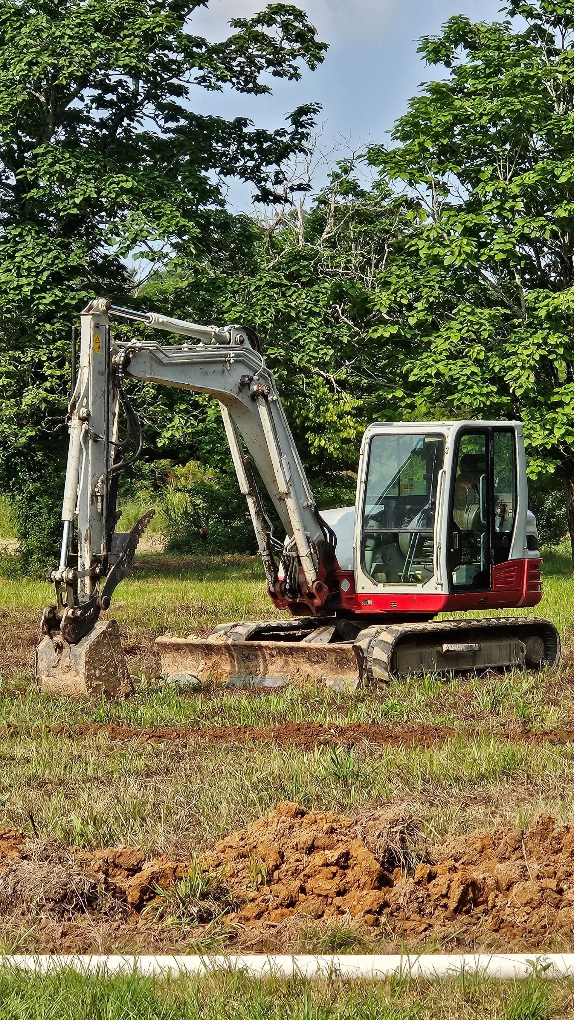 A white and red mini-excavator sits on a field of partially cleared land in front of green trees.