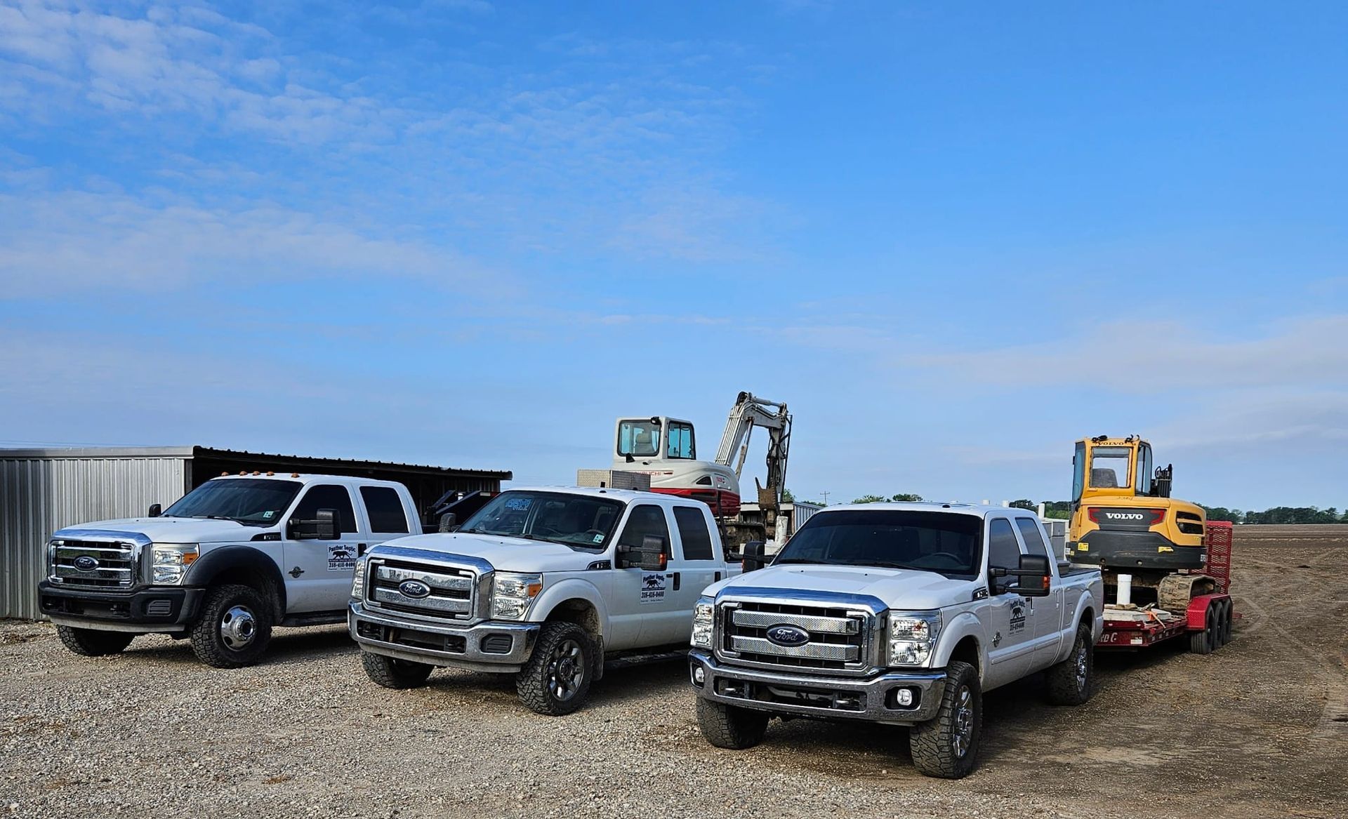 Heavy construction equipment, including a loader and two excavators, works on a cleared, dirt-covered site near trees.
