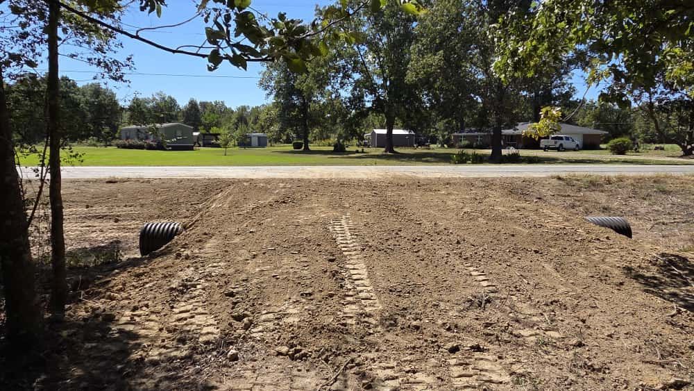 A rural driveway under construction, featuring a freshly graded dirt path with drainage pipes installed at the entrance.