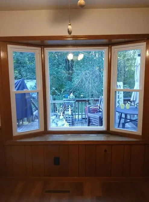 Bay window overlooking a deck and trees, with wood paneling and indoor lights visible inside.