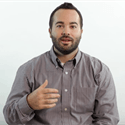 A person with a beard, wearing a grey button-down shirt, gesturing while speaking against a plain white background.