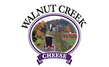 Logo for Walnut Creek Cheese featuring a circular scene of a horse-drawn carriage on a rural road with trees and a house.