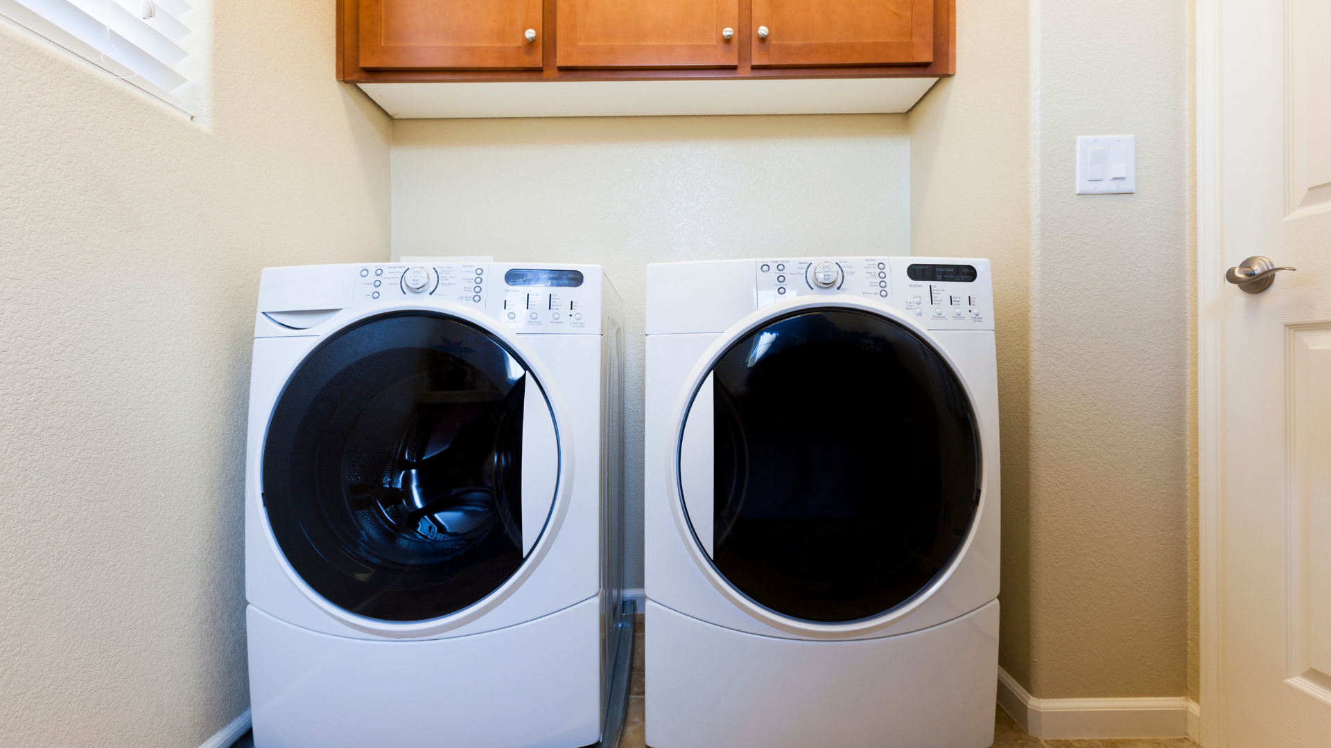 Two white washing machines in a laundry room, beneath wooden cabinets.