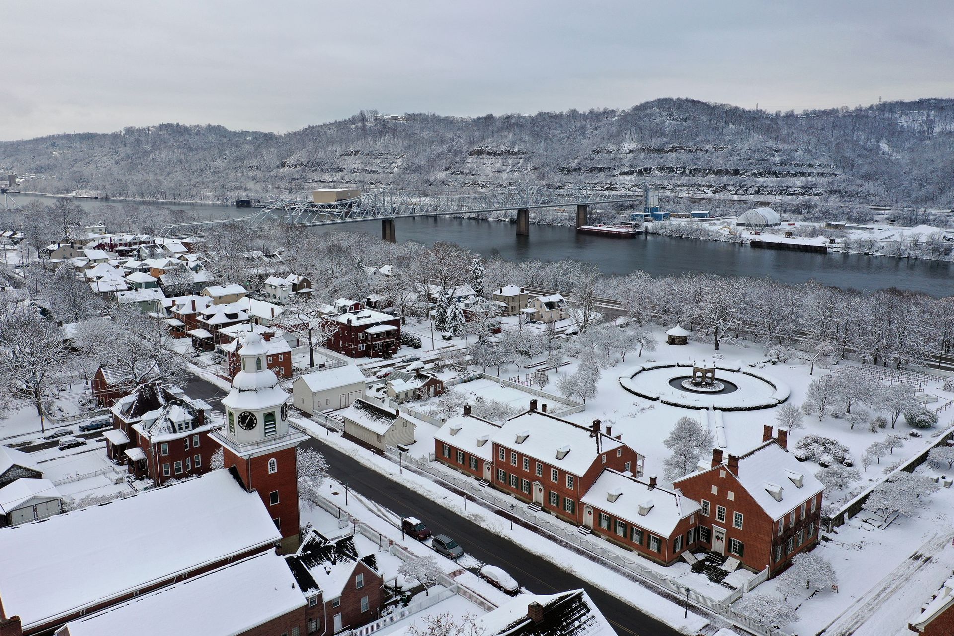 Snowy cityscape with red-roofed buildings by a river, viewed from above with hills in the background