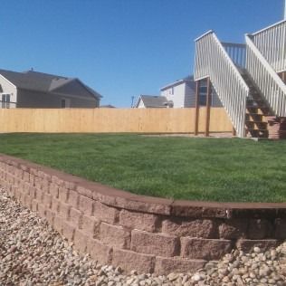 Backyard with green lawn, tan fence, stone retaining wall, and house stairs under a clear blue sky