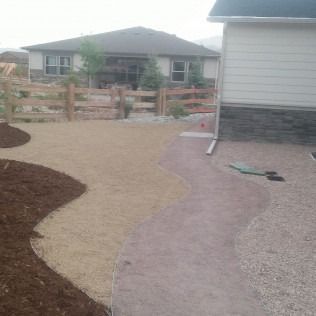 Curved gravel path beside a house, bordered by mulch and a wooden fence in a residential yard.