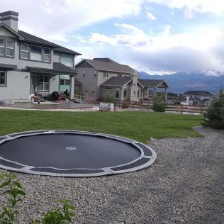 Backyard with a round trampoline, gravel path, green lawn, and suburban houses under a cloudy sky