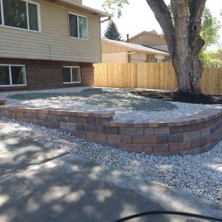 Raised brick planter with gravel in a backyard beside a house and wooden fence.