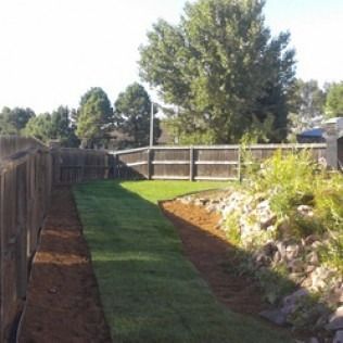 Backyard path with green grass strip, wooden fence, and rocks beside a raised garden bed