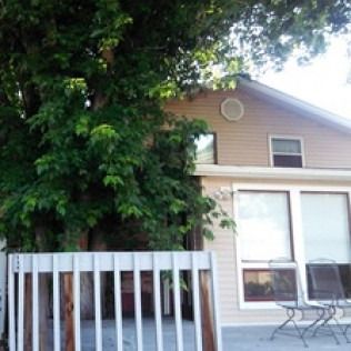 Small tan house with a white porch railing, large front window, and a leafy tree covering the left side.