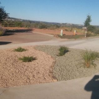 Curved desert road with gravel landscaping, sparse shrubs, and distant hills under a clear sky