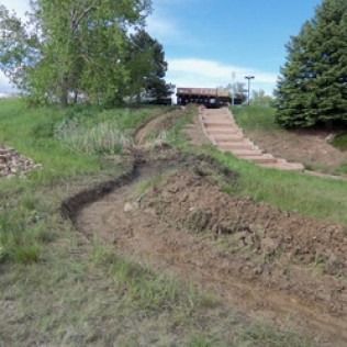 Rutted dirt slope beside concrete steps, leading up to a road in a grassy park area.