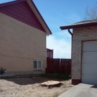 A narrow dirt alley between two beige houses, with a red fence and blue sky in the background.