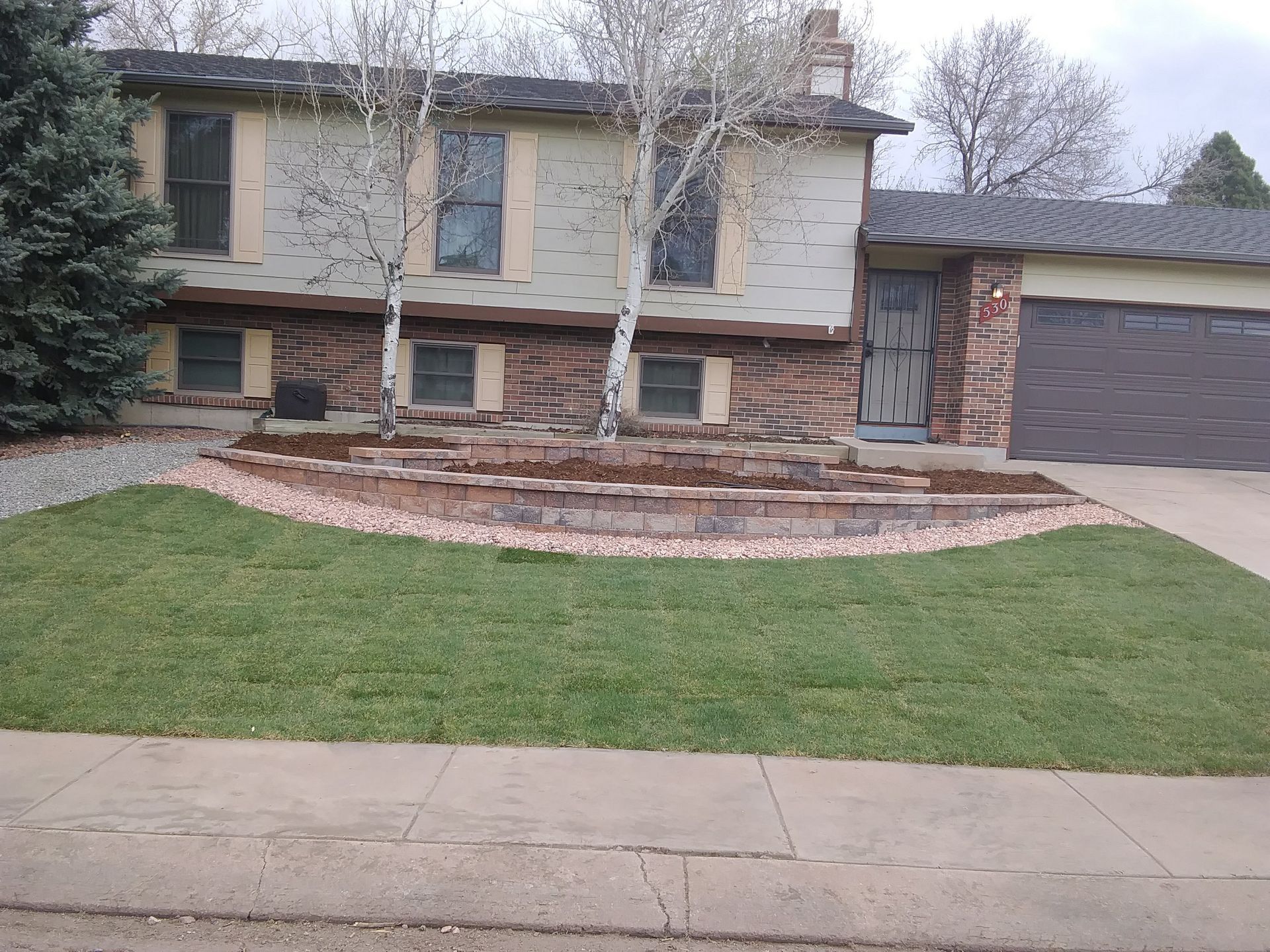 Two-story suburban house with brick facade, double garage, and front lawn with leafless trees.