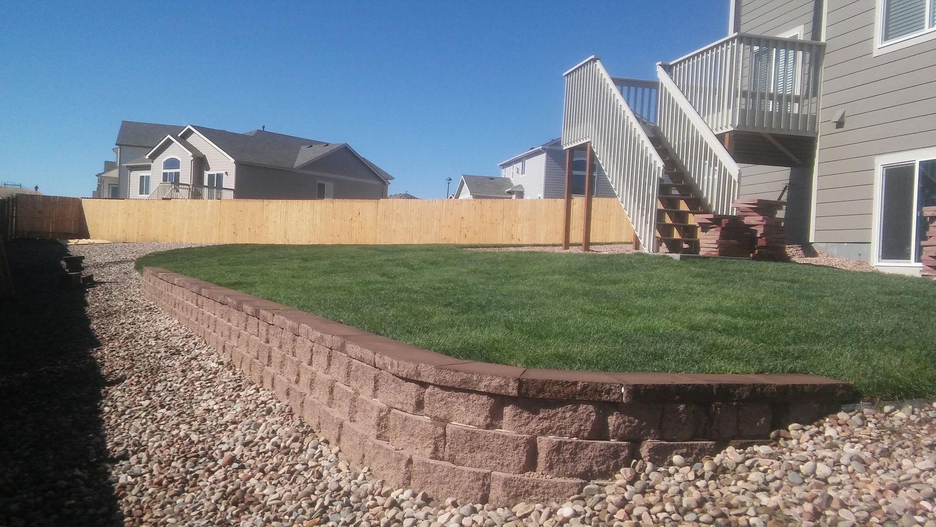 Backyard with brick retaining wall, green lawn, rock border, and a house with exterior stairs