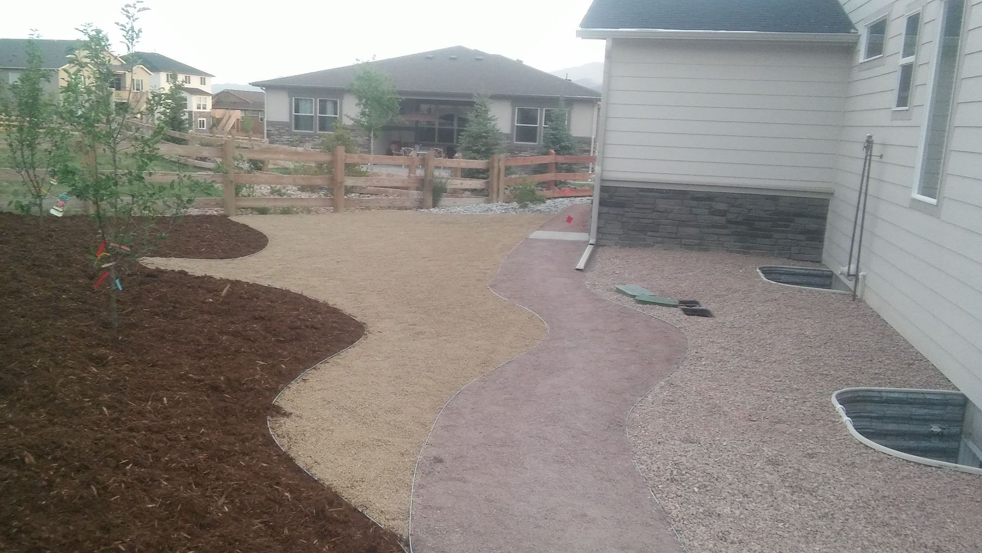 Landscaped backyard with a curved gravel path, mulch beds, wooden fence, and a house exterior.