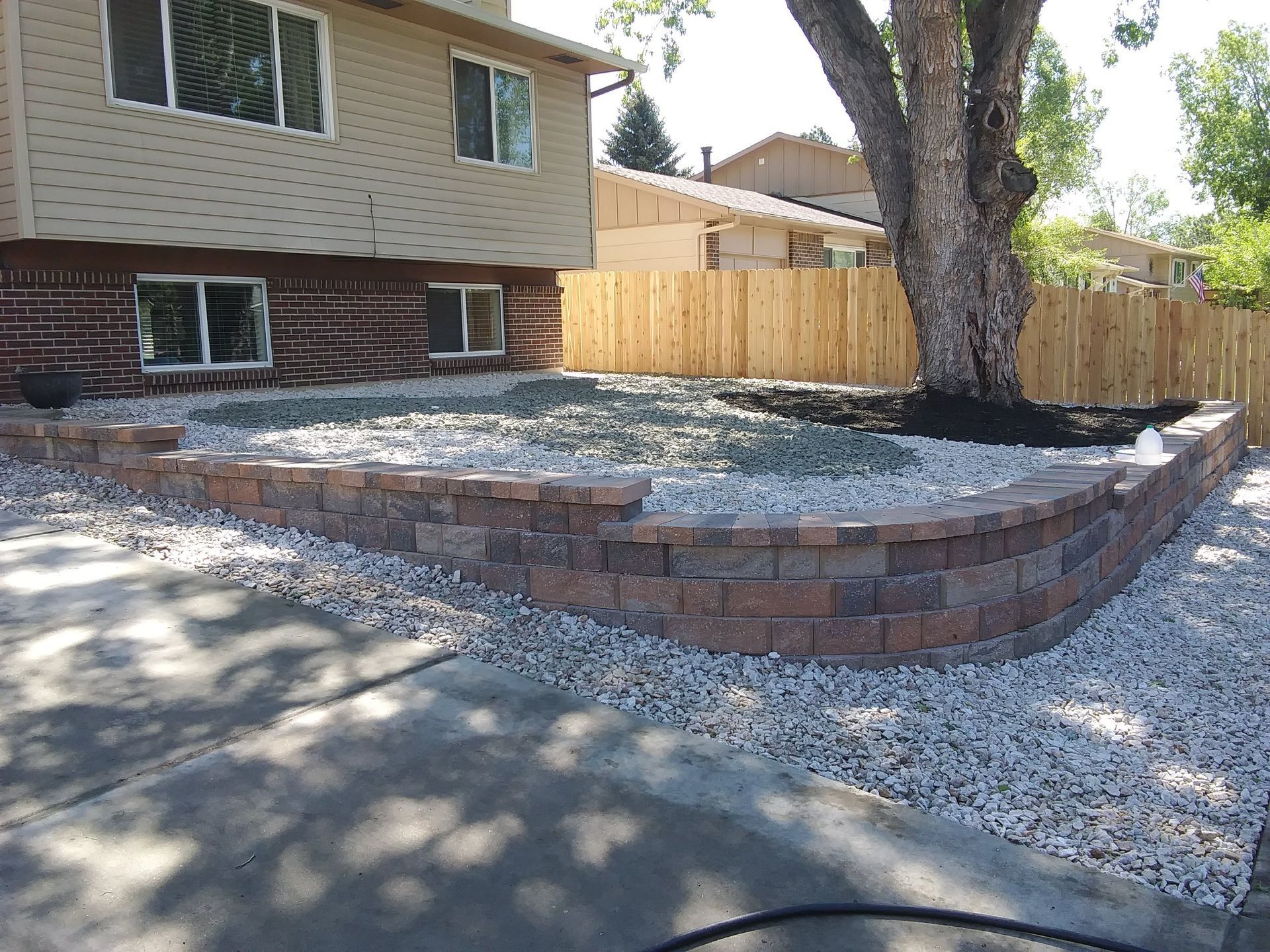 Raised backyard patio with brick retaining wall, gravel ground cover, and a large tree beside a house
