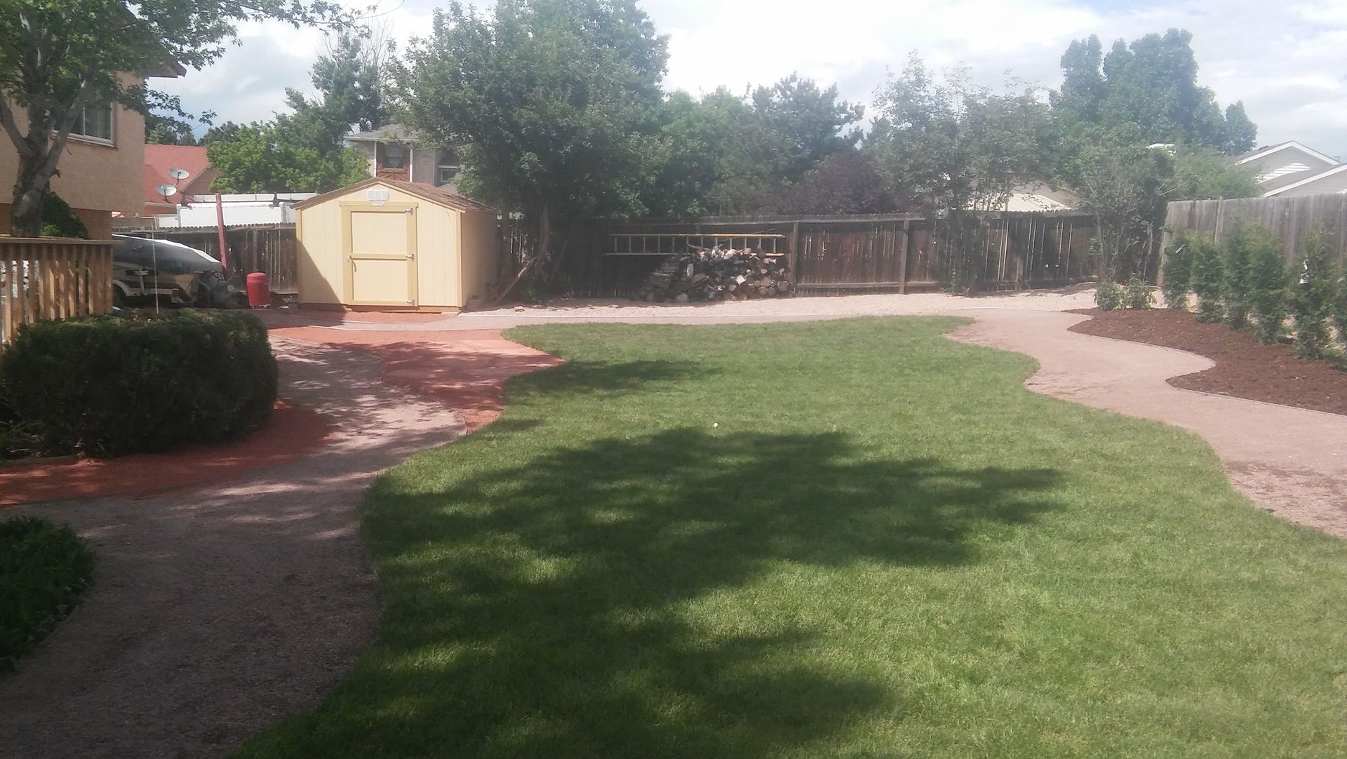 Sunny backyard with a green lawn, curved gravel paths, trees, and a beige shed near the fence
