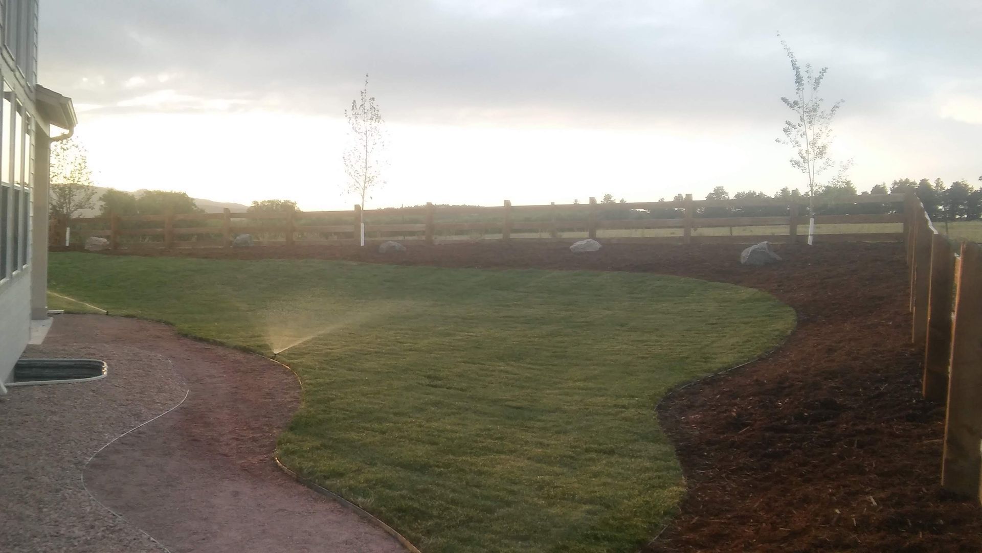 Curved green lawn beside a house, with a gravel path and open field at sunset.
