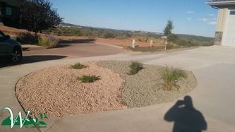 Desert landscaping with gravel mounds and small shrubs beside a driveway, with a building and road in the background.