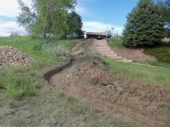 Grassy hill with muddy erosion path, stone steps, and trees under a cloudy sky