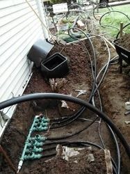 Garden bed with black hoses and irrigation tubing beside a house siding, over mulch and soil.