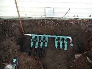 Exposed underground plumbing manifold with green valves and black pipes against a house foundation in a dug trench