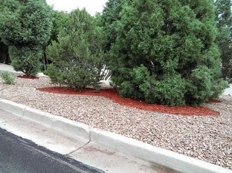 Landscaped curb with green shrubs, red mulch, and gravel beside a paved road