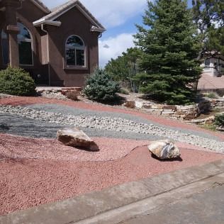 Front yard with red gravel landscaping, large rocks, shrubs, and a brown house under a blue sky