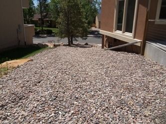 Rock-covered landscape strip beside beige apartment buildings and a tree in a sunny courtyard