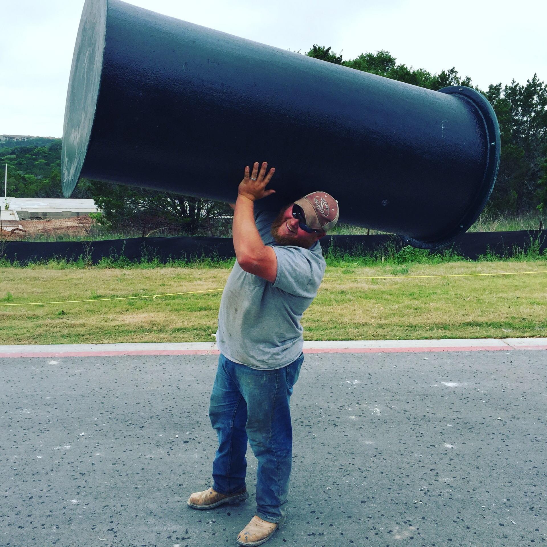 Man lifting large, black cylindrical pipe outdoors. Gray shirt, jeans.