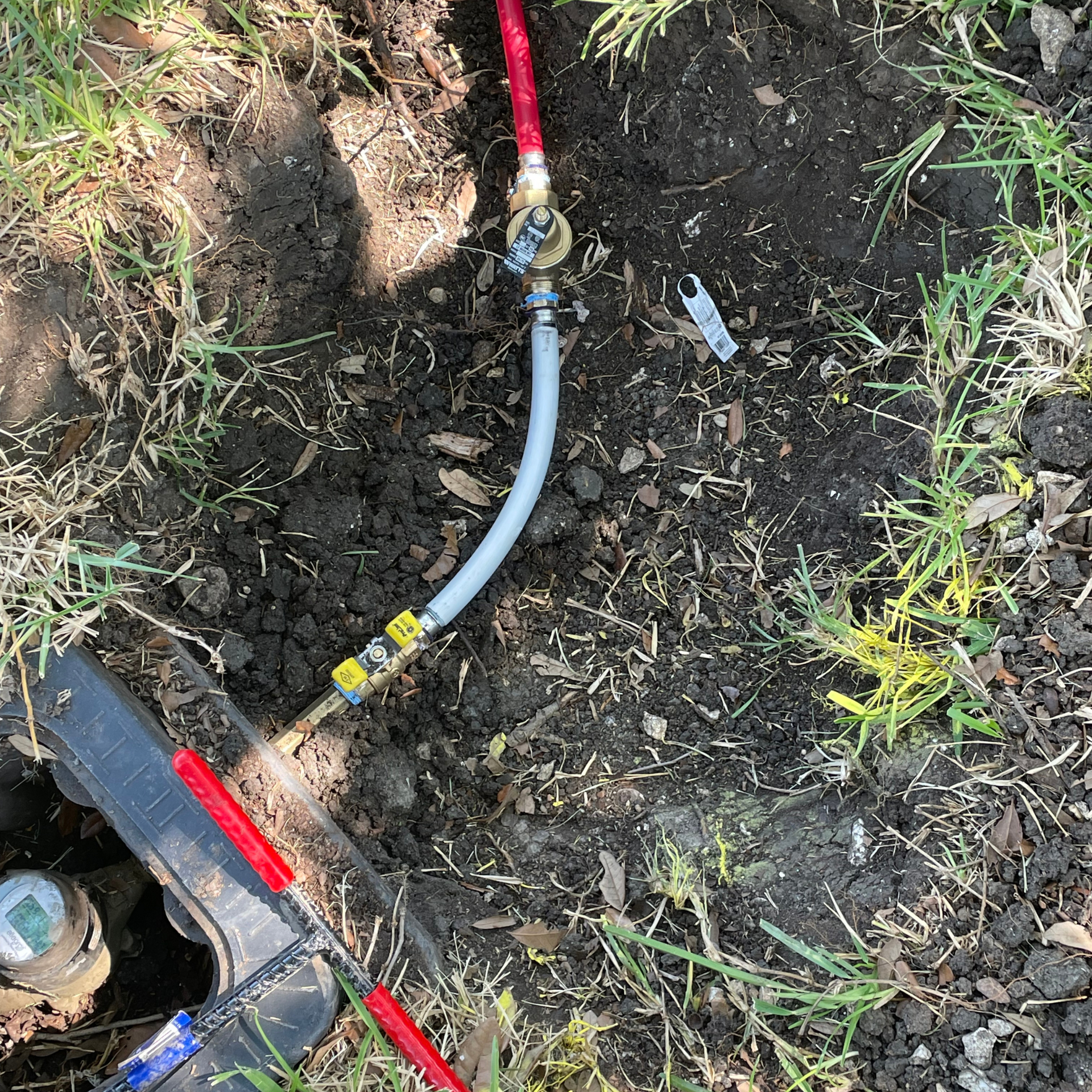 Plumbing connections in a dirt patch; red and gray hoses, brass fixtures, and a water meter.