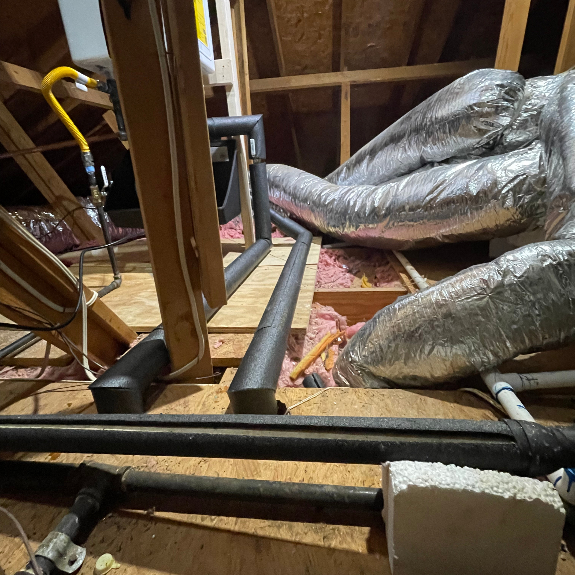 Attic with wooden beams, insulation, ductwork, and pipes covered in black insulation.