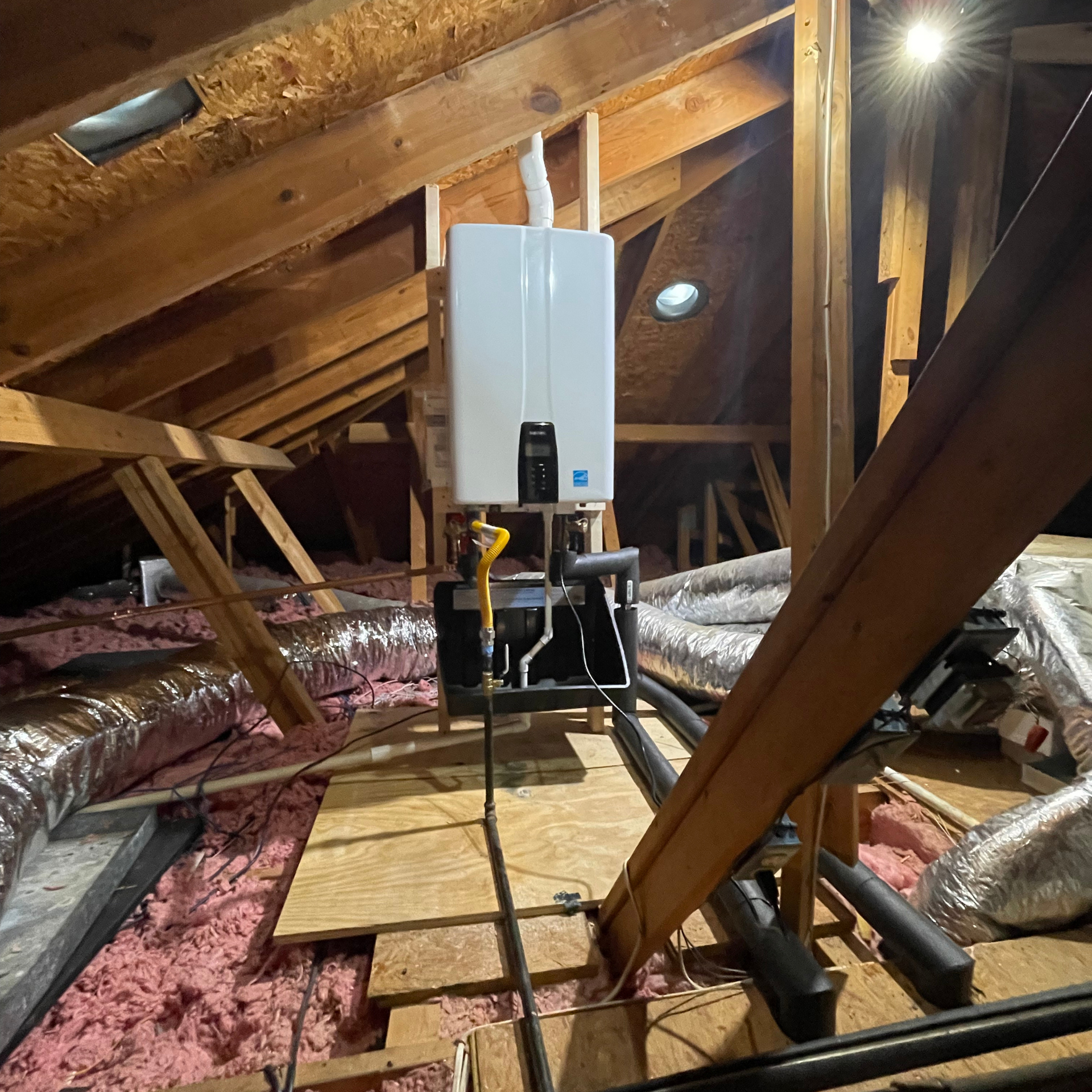 Tankless water heater installed in an attic. It is white, with vents and pipes, on a wooden platform among insulation.