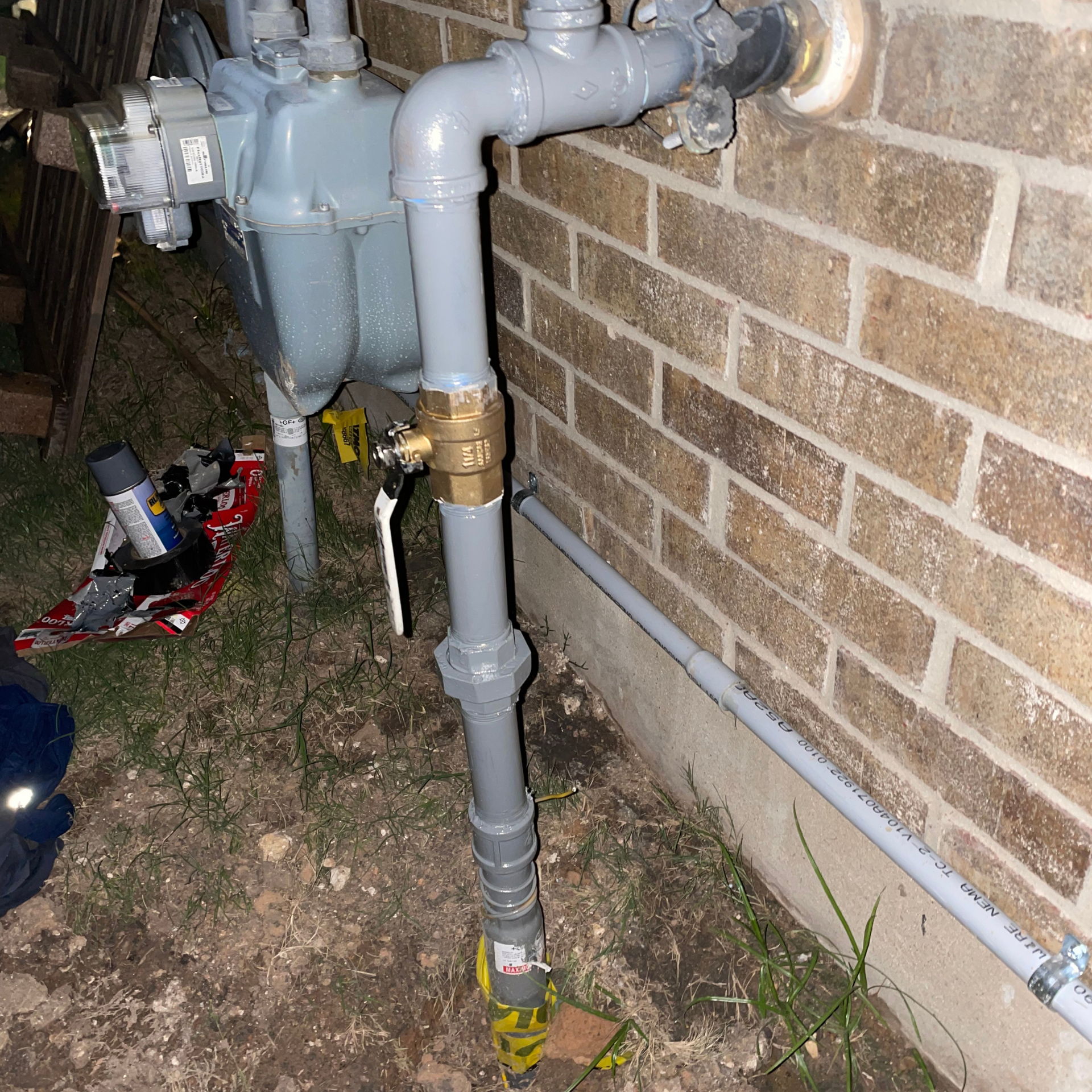 Gas meter and piping attached to a brick wall. Gray pipes and brass fittings are visible.