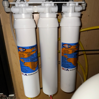 Three white water filters with blue and orange labels, mounted on a wooden surface, connected by tubing.