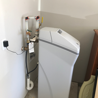Water softener with connected pipes and power outlet against a white wall.