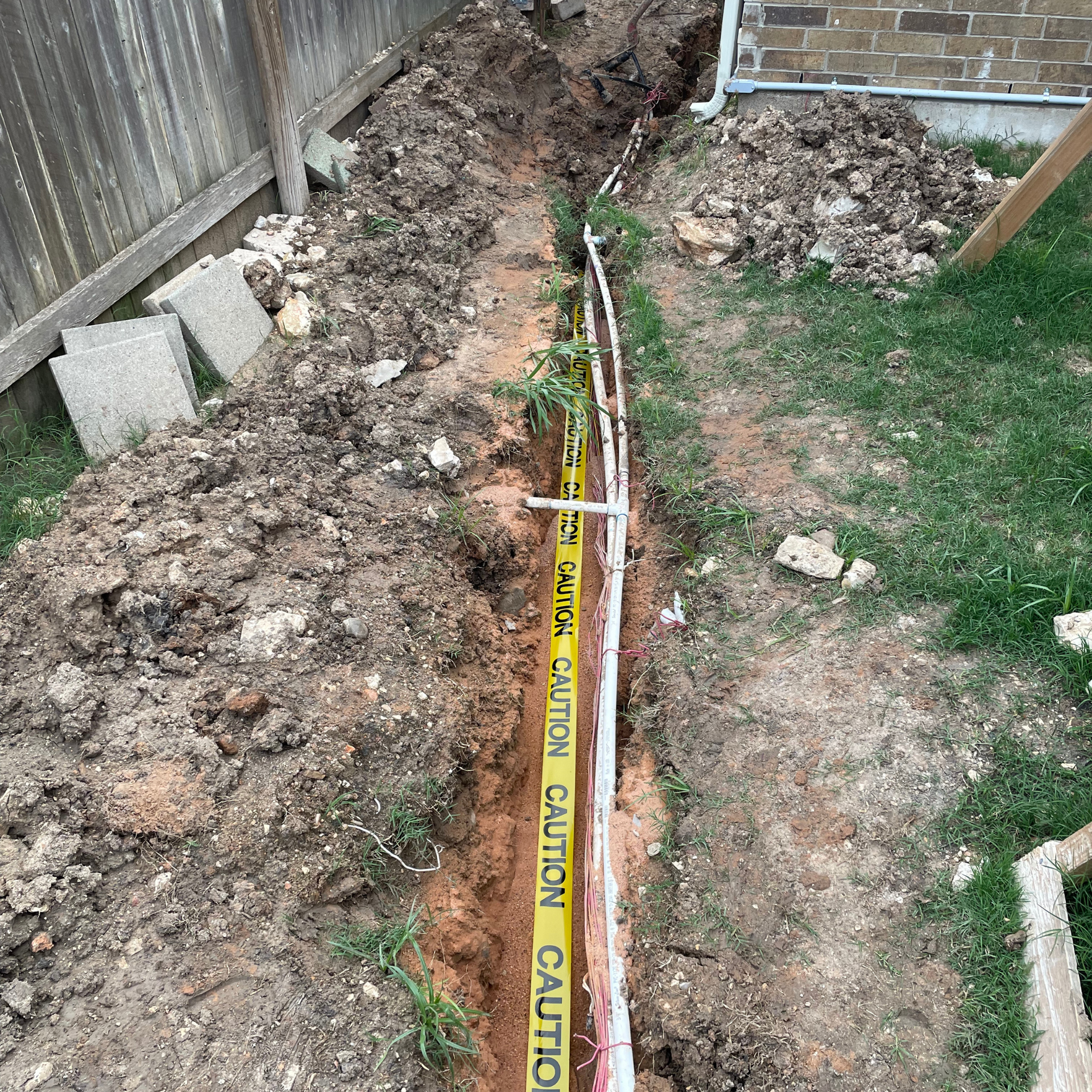 Trench dug in yard with yellow and white pipes. Caution tape is visible.