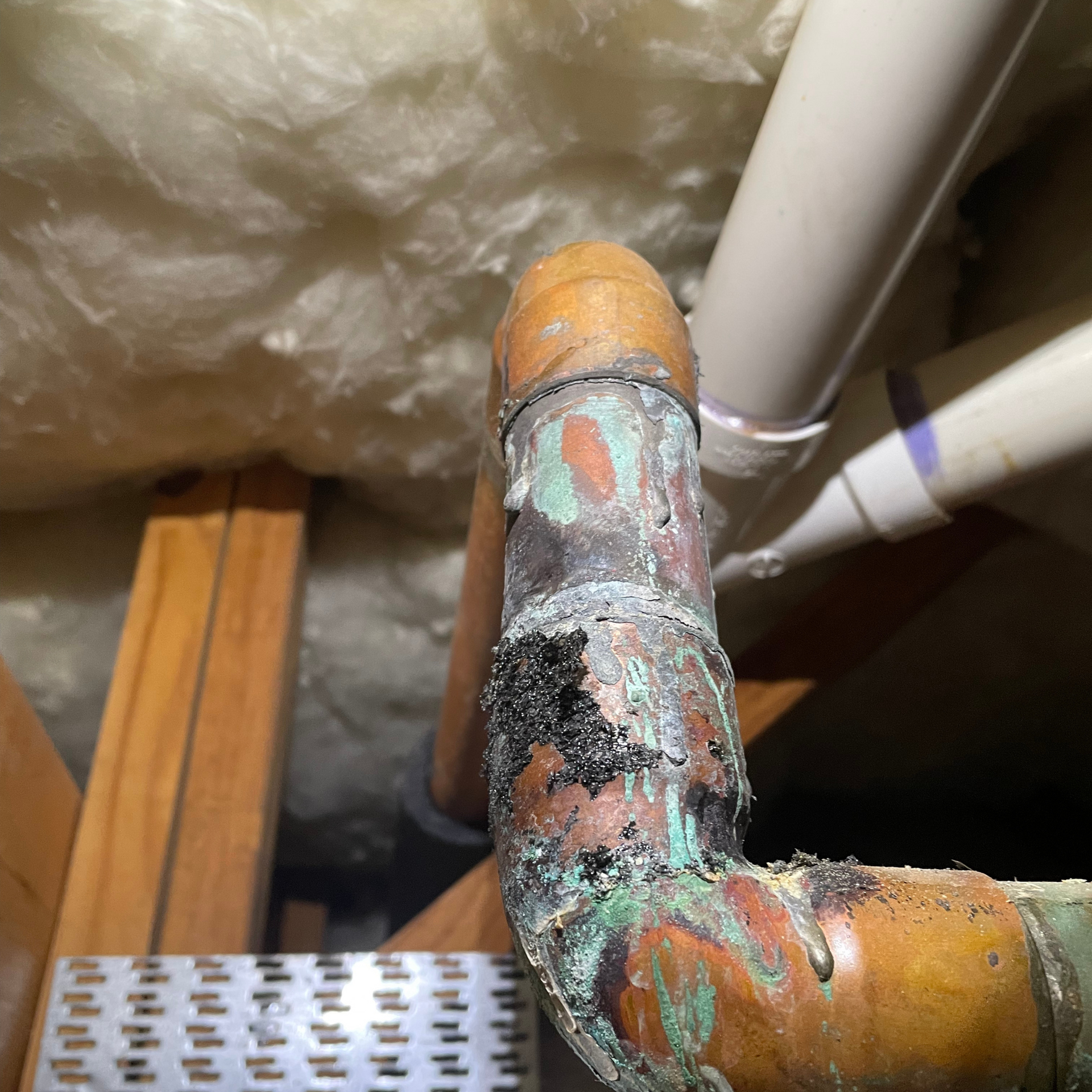 Corroded copper pipe with green and black oxidation in an attic, near insulation and a wooden frame.