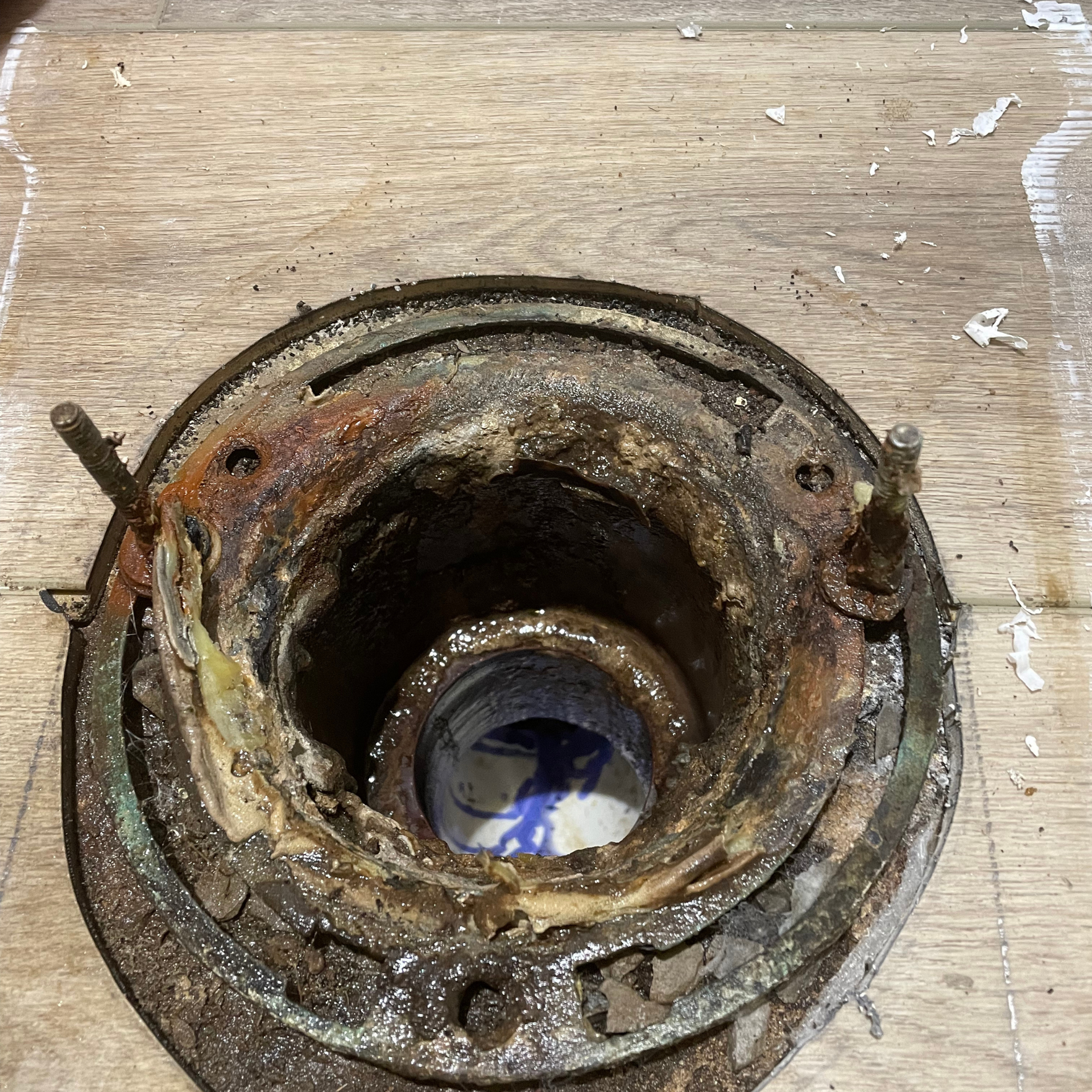 Close-up of a corroded toilet flange and waste pipe, surrounded by wooden flooring.