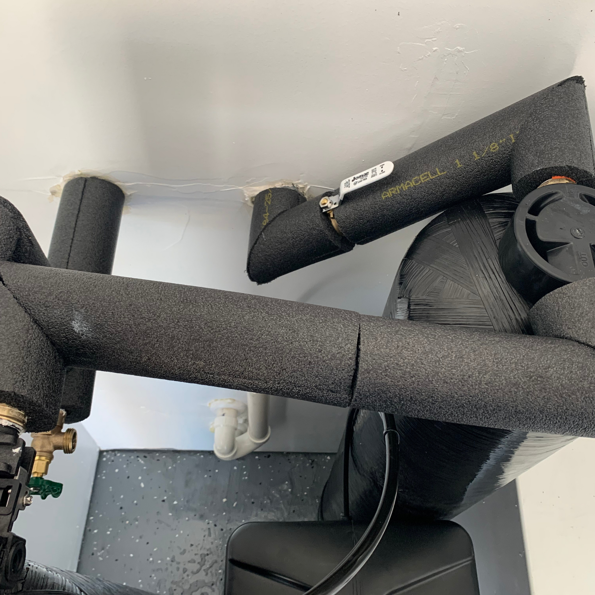 Black insulated pipes connected to a pump and valves, against a white wall and dark floor.