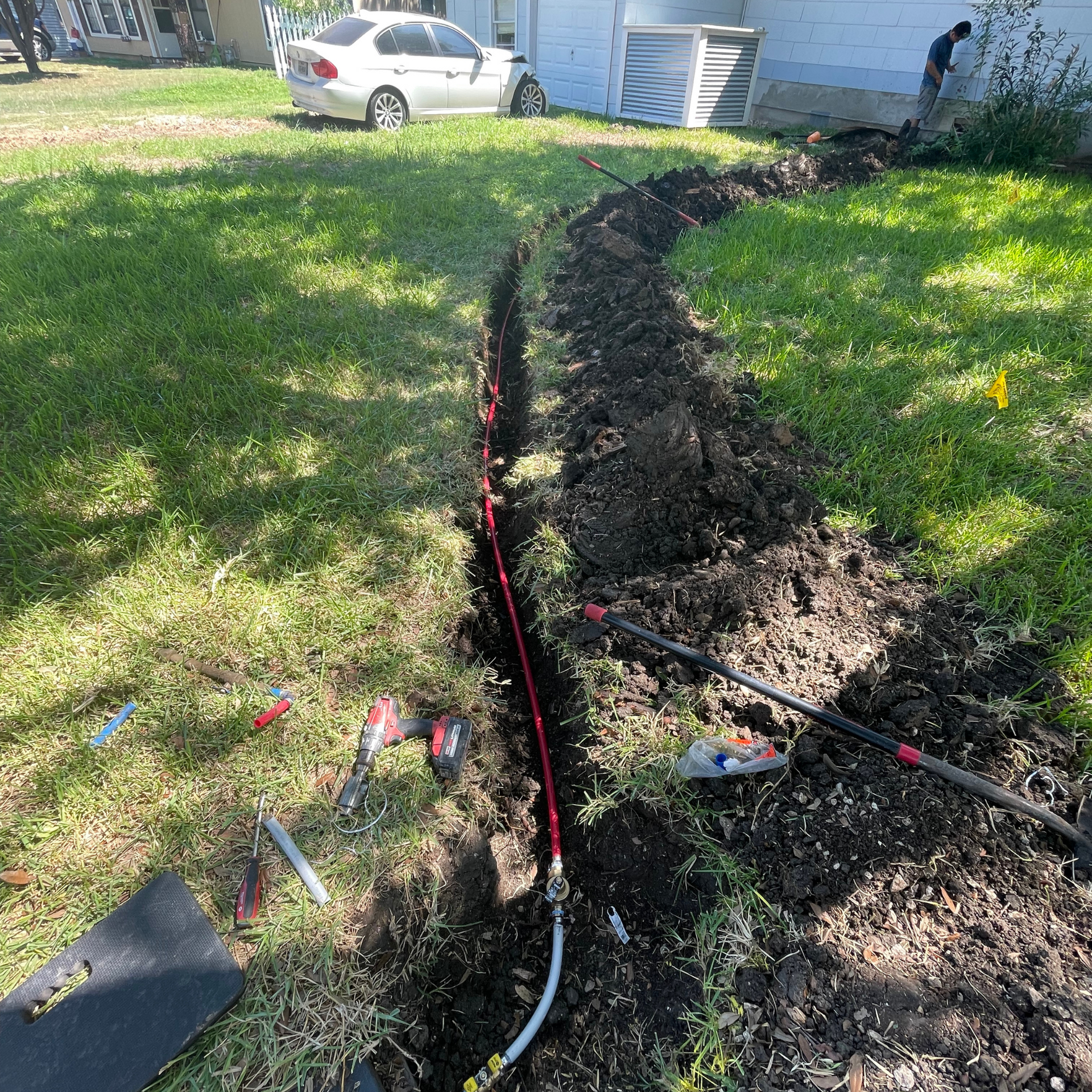 Trench in grass with a red gas line, tools, and a car in the background.
