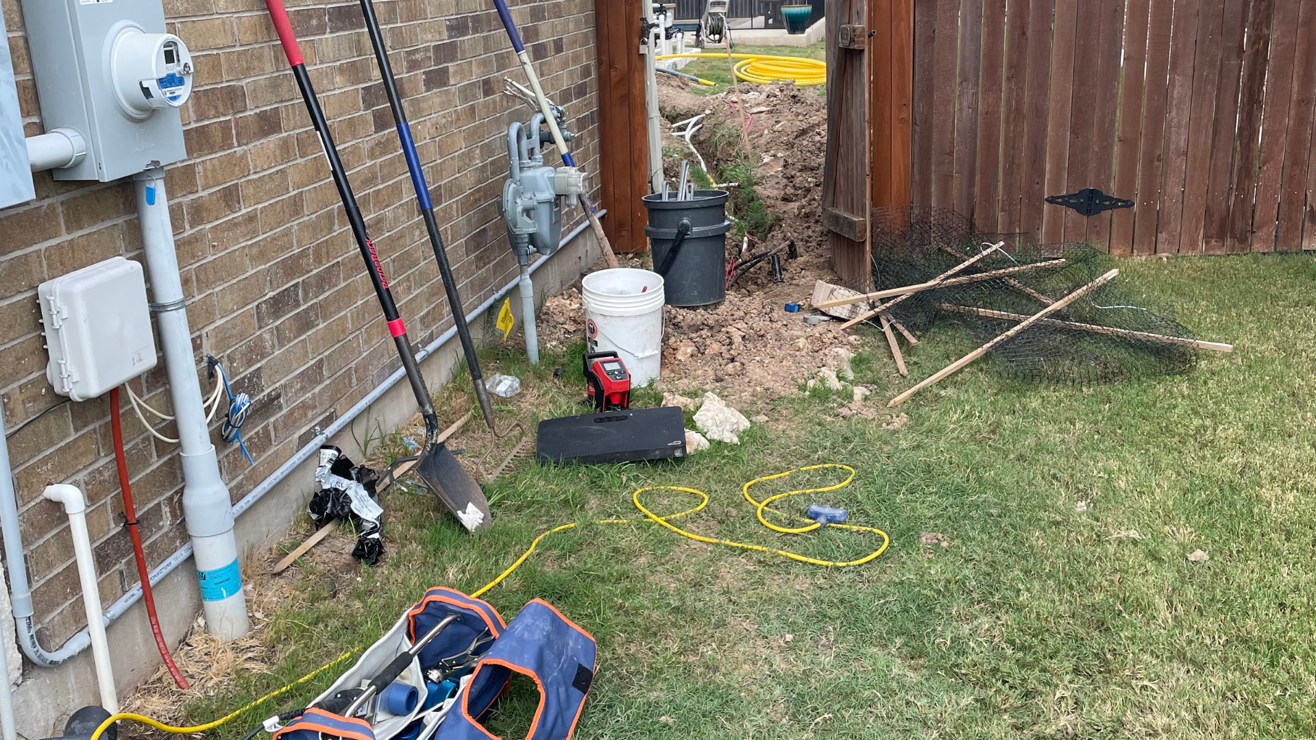 Tools and equipment on grass next to a brick wall and wooden fence.