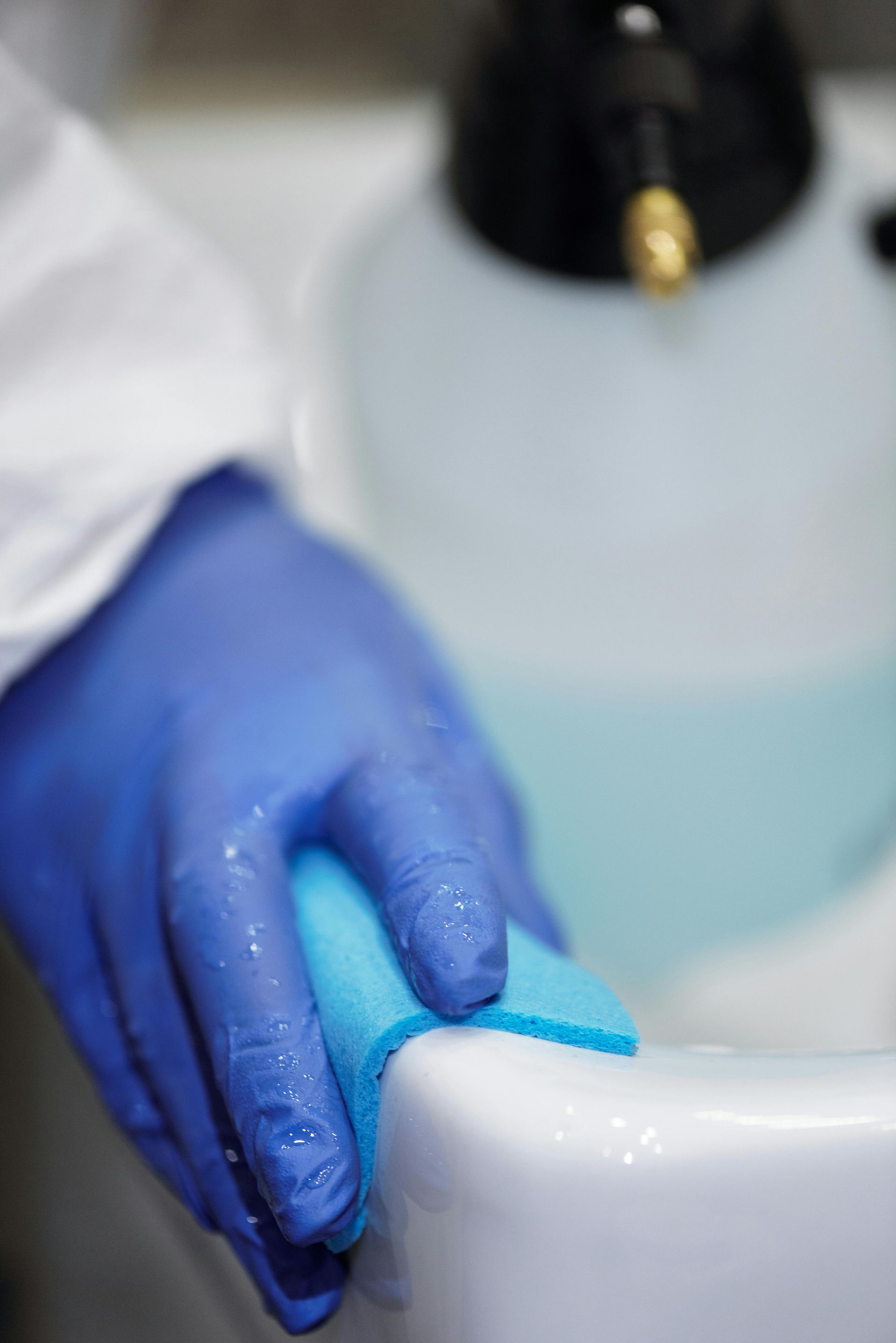 Hand in blue glove wiping a white surface with a blue sponge; spray bottle in the background.