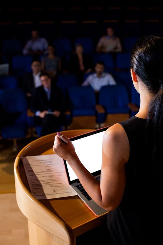 Orador en el podio dirigiéndose a una audiencia en un aula de conferencias con poca luz.