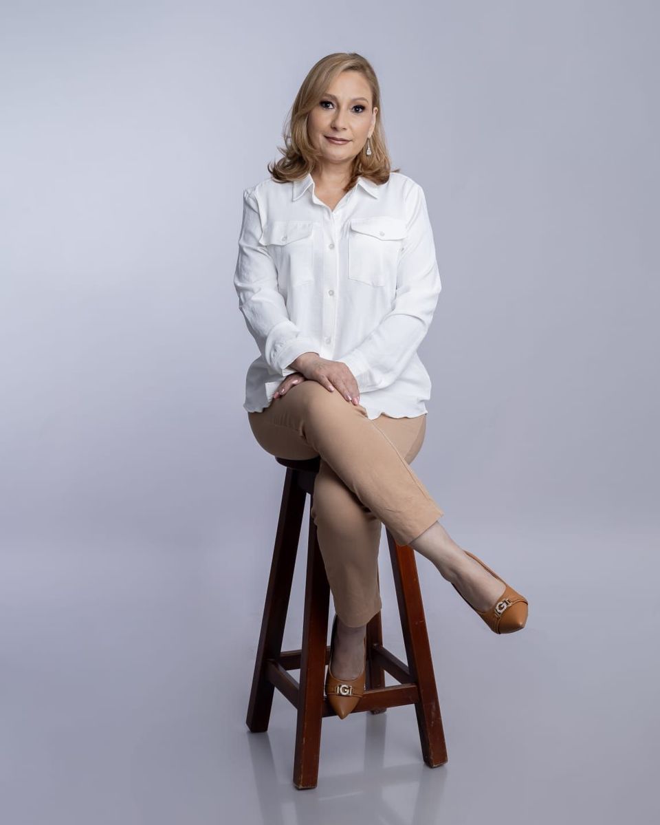 Mujer sentada en un taburete de madera, con camisa blanca y tacones color canela, frente a un fondo de estudio gris.