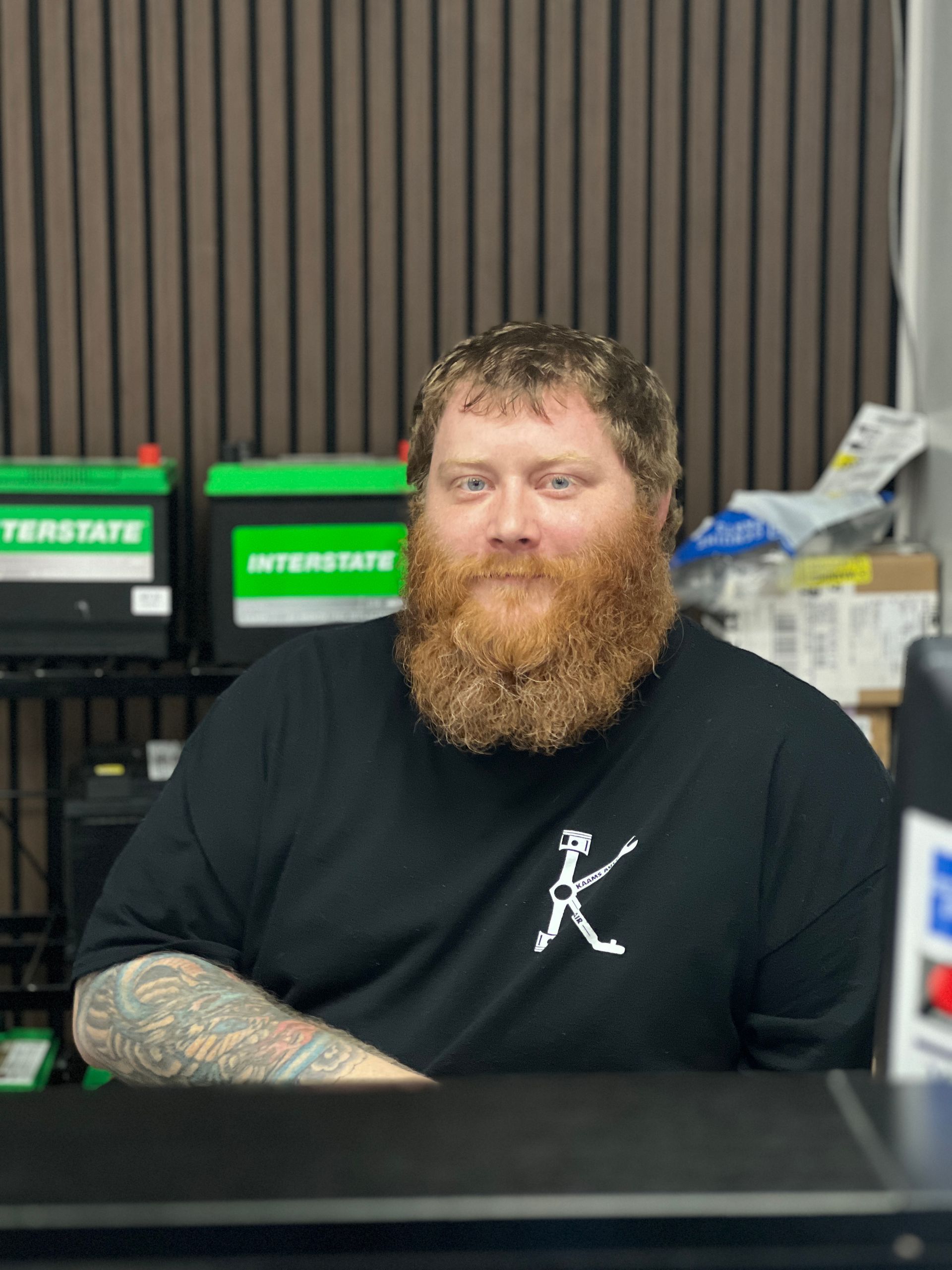 Man with red beard, tattoo on arm, in front of Interstate batteries. Black shirt with white